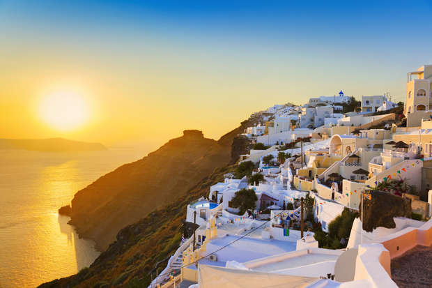 White buildings perched on a hillside reflect golden sunlight, overlooking a vast, calm sea during sunset. The blue sky transitions smoothly to orange, enhancing the picturesque coastal vista.