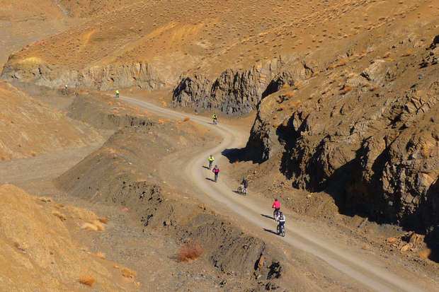 Bicyclists ride down a winding dirt road, surrounded by rugged, arid hills with sparse vegetation, creating a sense of adventure in an isolated desert landscape.