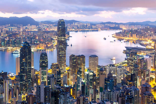 Skyscrapers illuminate the bustling cityscape with diverse buildings towering over the water. The harbor reflects city lights as dusk transitions to night, with mountains silhouetted in the background.