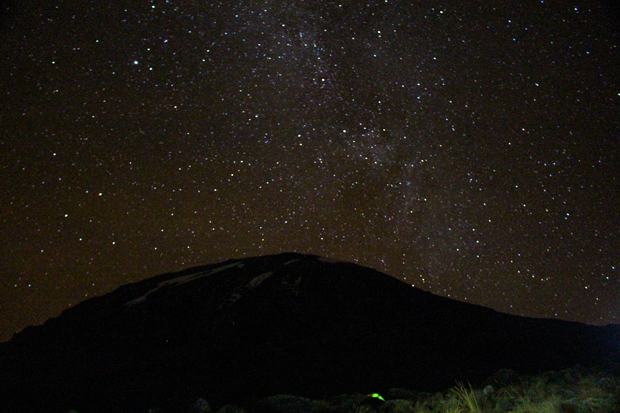 A silhouetted mountain rests under a star-filled night sky. Distant stars form a dense, bright cluster, possibly the Milky Way, above. Grassy terrain and rocks are faintly visible below.