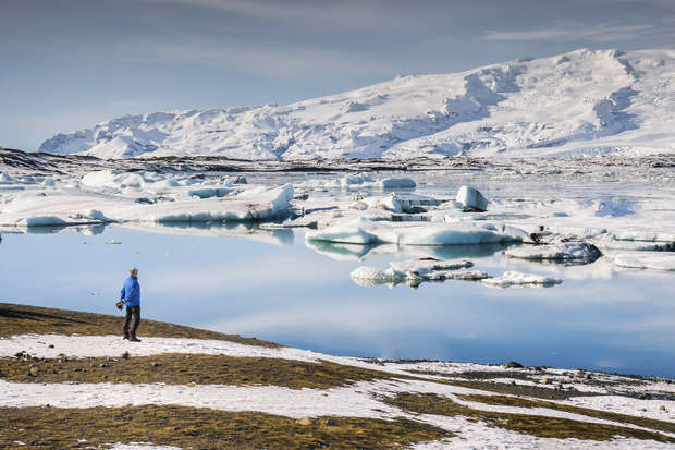 A person stands on snow-dusted ground, looking at icebergs floating in a calm, reflective body of water, with a snow-covered mountain range in the background under a clear sky.
