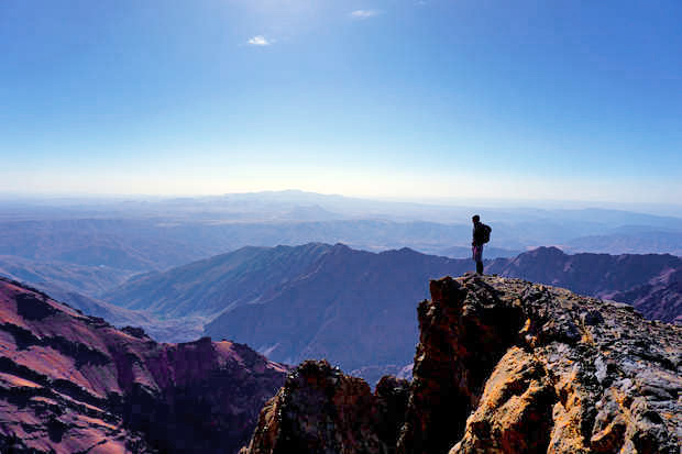 A lone hiker stands atop a rocky cliff, gazing at a vast, layered mountain landscape under a clear blue sky.