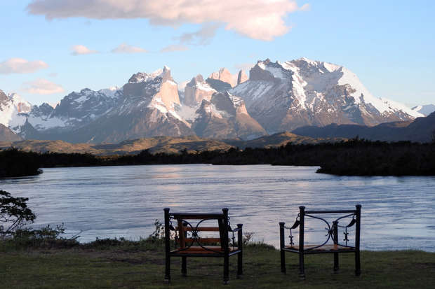Two wooden chairs facing a tranquil river, with a backdrop of rugged snow-capped mountains under a partly cloudy blue sky. The setting is serene and picturesque.