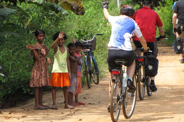 Cyclists ride along a dirt path, waving at four children standing nearby, surrounded by lush greenery and parked bicycles, under a partially shaded area.