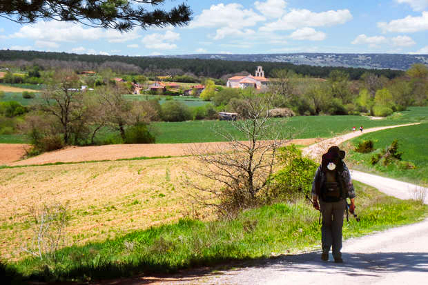 A solitary hiker walks along a rural path wearing a backpack and hat, with fields and a village with a church visible under a partly cloudy sky in the background.