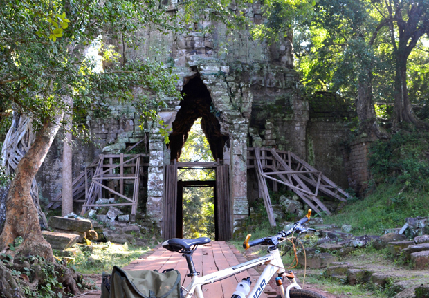 Bicycle rests on a wooden path leading to a weathered stone archway, partially supported by wooden beams; surrounded by lush trees and greenery in a historical ruin setting.