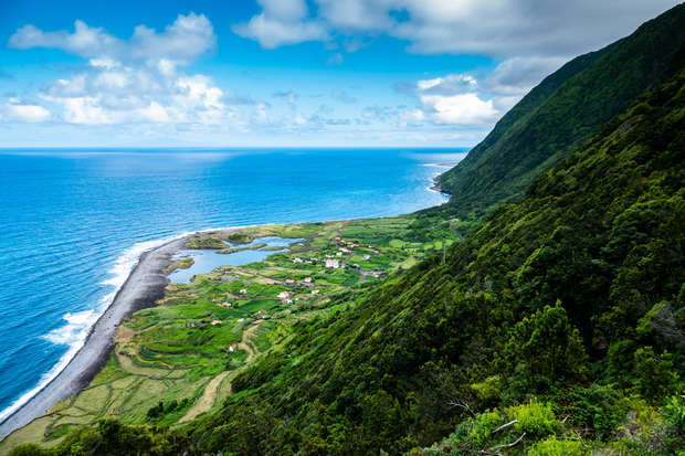 A lush green hillside slopes towards a coastal village, bordered by the ocean; scattered clouds dot the blue sky, creating a serene landscape blending land and sea.