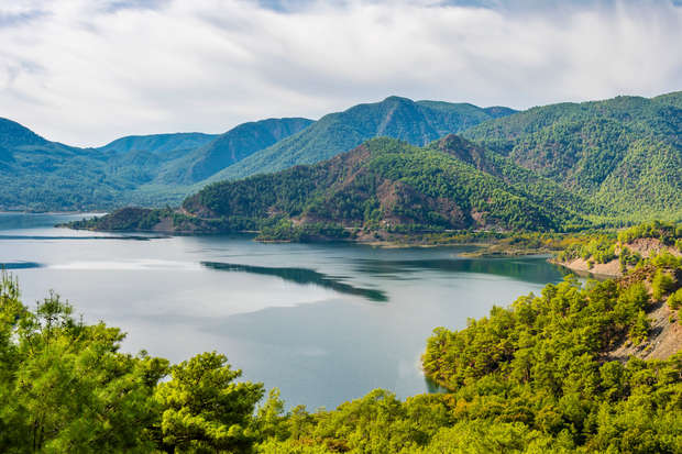 A tranquil lake reflects surrounding lush green forests and rolling mountains, under a partially cloudy sky, creating a serene natural landscape.