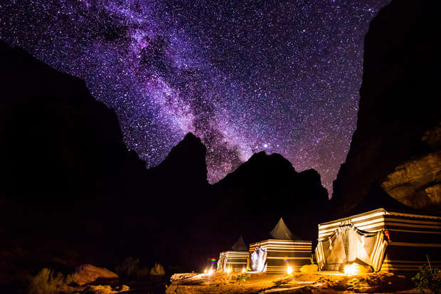 Tents are illuminated with warm lights at night, set against dramatic silhouetted mountains and a vivid, star-filled sky displaying the Milky Way.