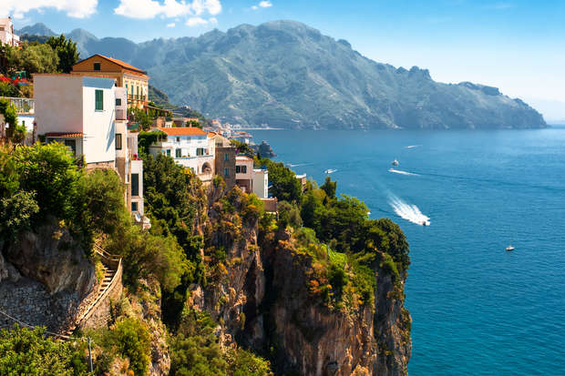Cliffside houses perch atop lush, rocky terrain overlooking a calm, expansive blue sea with boats creating white trails, set against distant, hazy mountains under a clear, sunny sky.