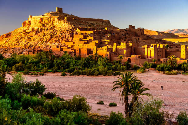 Ancient fortress sits atop a sunlit hill, surrounded by desert landscape and lush greenery. A clear sky and distant mountains frame the scene, enhancing the historical, serene atmosphere.