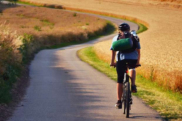 A cyclist rides along a winding rural road, surrounded by golden fields and tall grass, carrying a large green backpack under a sunny sky.