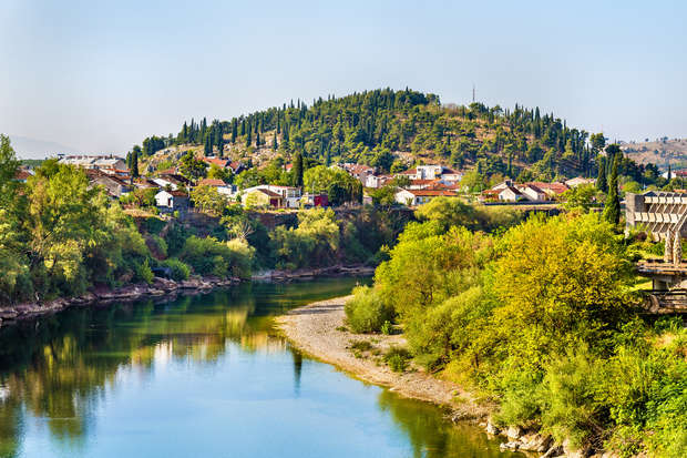A serene river flows through a lush landscape, bordered by dense greenery. Houses dot the hill in the background, surrounded by tall trees under a clear, blue sky.