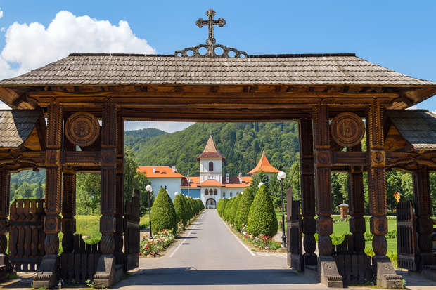 A large, intricately carved wooden gate frames a pathway lined with manicured shrubs, leading to a distant church with a red-roofed tower, set against a backdrop of lush, forested hills.