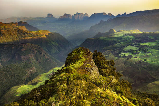 Rugged mountain peak covered in green vegetation under soft morning light, overlooking vast valleys and distant, hazy mountain ranges, creating a serene and expansive landscape view.