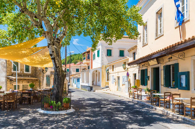 A large tree provides shade over wooden chairs and tables on a sunlit street. Colorful buildings with shutters line the road, and a Greek flag is displayed.