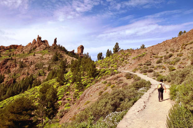 Hiker walks along a winding dirt path amidst rocky, green-vegetated hills and towering rock formations under a partly cloudy blue sky.