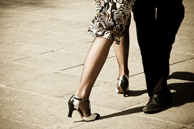 Two elegantly dressed legs perform a dance on a sunlit, tiled pavement; the woman's legs are in patterned stockings and heels, while the man's legs are in dark trousers and shoes.