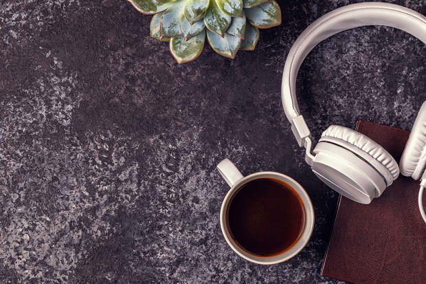 White headphones rest on a closed book beside a coffee mug on a textured dark surface, with a green succulent plant nearby, creating a relaxed, cozy atmosphere.