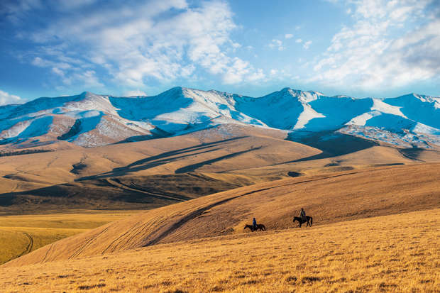 Two horseback riders traverse golden grassy hills, backed by snow-capped mountains under a partly cloudy blue sky.