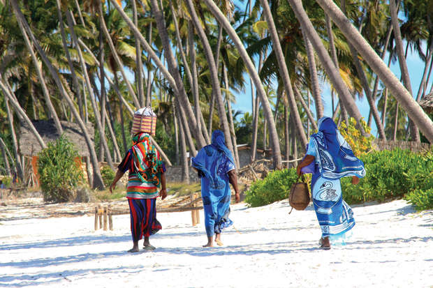 Three people walk along a sandy beach, wearing colorful garments. One carries items on their head. Tall palm trees and lush greenery surround the area, evoking a tropical setting.