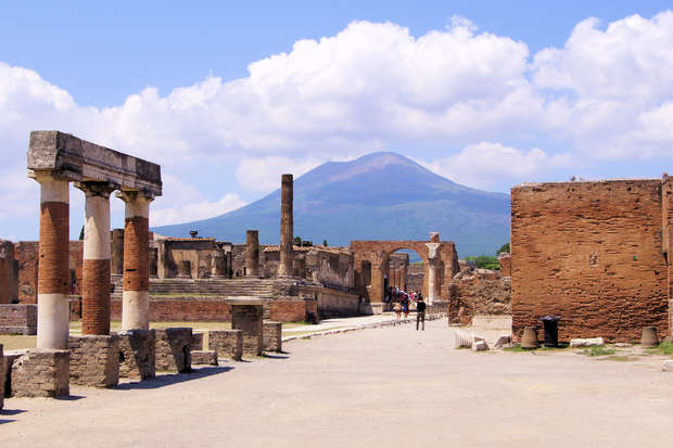 Ancient stone columns and ruins stand under a blue sky, with Mount Vesuvius in the background. Visitors walk along the pathway, exploring the historic site amidst partially cloudy weather.