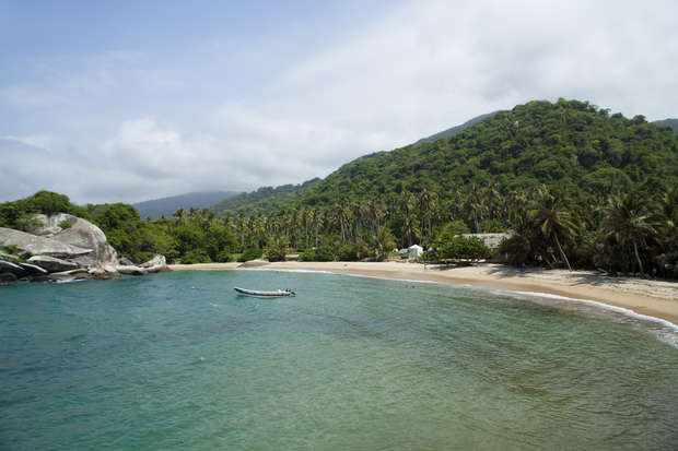 A small boat floats on clear turquoise water, near a sandy beach lined with lush palm trees, backed by a forested hillside under a partly cloudy sky.