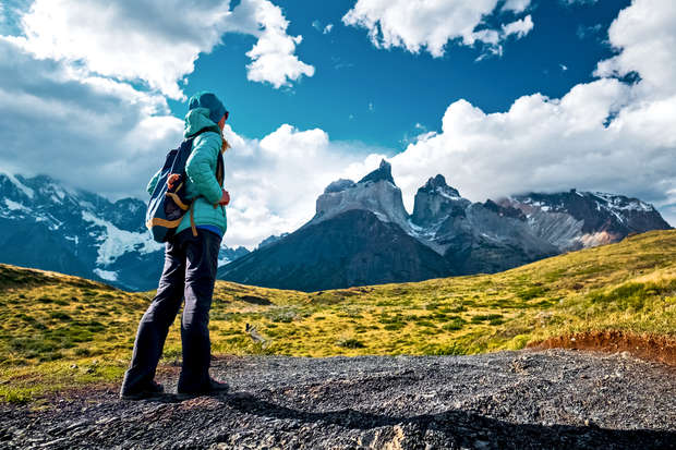 A person wearing outdoor gear stands on a rocky path, gazing at distant snow-capped mountains under a partly cloudy sky in a vast, grassy landscape.