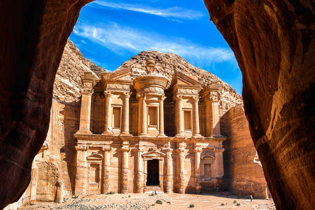 A grand, ancient stone building with carved columns and intricate facades stands under a vivid blue sky, viewed through a natural rock archway, surrounded by a desert landscape.