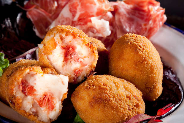 Golden, fried croquettes rest on a plate, some cut open to reveal creamy filling with bits of red meat, accompanied by thinly sliced cured ham in the background.