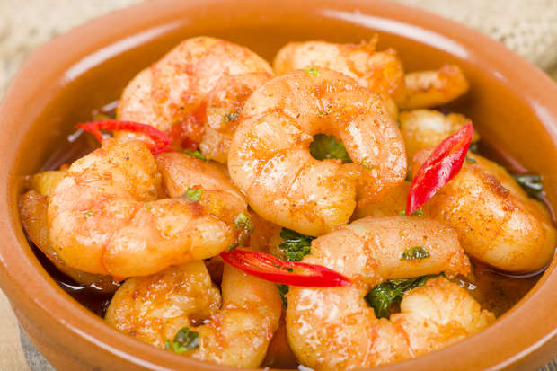 Shrimp are cooked and seasoned, garnished with sliced red chili peppers and herbs, served in a brown, rustic ceramic bowl, suggesting a spicy seafood dish ready to eat.