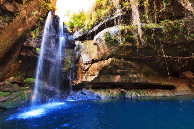 Waterfall cascading into a clear blue pool, surrounded by rugged rock formations and lush greenery, with sunlight illuminating the scene, creating a serene and natural atmosphere.