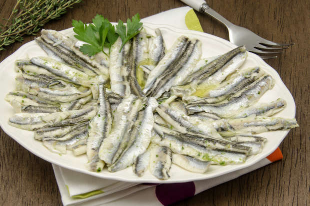 Marinated anchovies are arranged in a circular pattern on a white platter, garnished with parsley. A fork and sprig of herbs rest nearby on a wooden table.