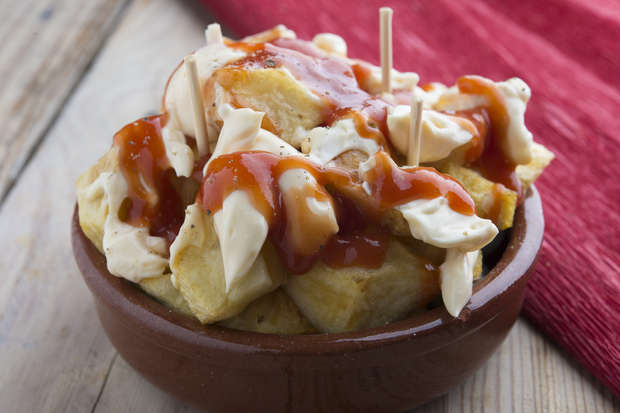 Golden fried potatoes topped with creamy white sauce and red sauce, served in a small brown bowl on a wooden table, next to a red cloth, with toothpicks inserted.