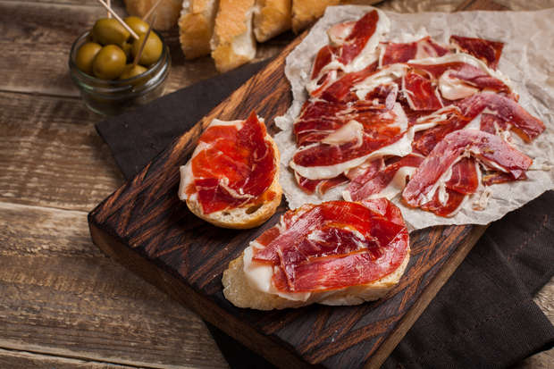 Slices of cured meat rest on bread and parchment, displayed on a wooden board. A jar of green olives with toothpicks and crusty bread are nearby on a rustic wooden table.