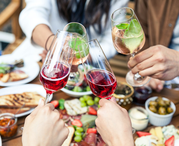 Hands clink wine glasses containing red and rose wine over a table filled with dishes, including bread, cheese, olives, and vegetables, in a social dining setting.