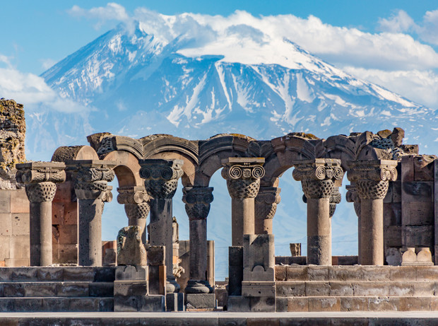 Stone columns stand in a row, showcasing intricate carvings, under a clear sky. Snow-capped mountains rise majestically in the background, highlighting the ancient architectural site.