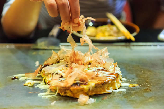 Hand sprinkles bonito flakes on okonomiyaki, a savory Japanese pancake, on a hot grill. Chopsticks and a plate of food are visible in the background.