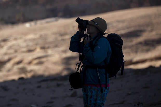 A person with a backpack and hat uses a camera, photographing a rugged, sunlit landscape. Shadows create a contrasting atmosphere against the bright, sandy terrain.