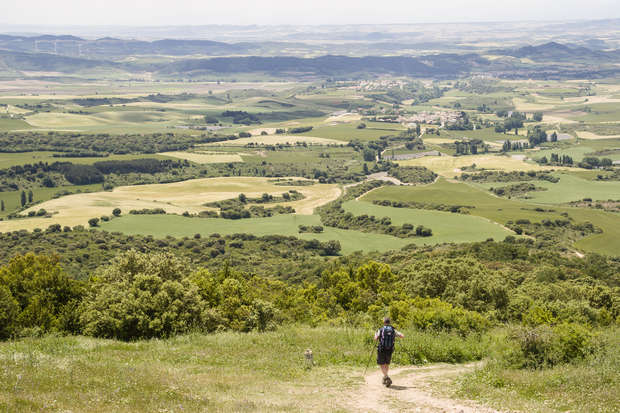 A person hikes along a dirt path through a lush, green landscape. Rolling hills and scattered trees stretch into the distance under a clear sky, with a village visible far away.