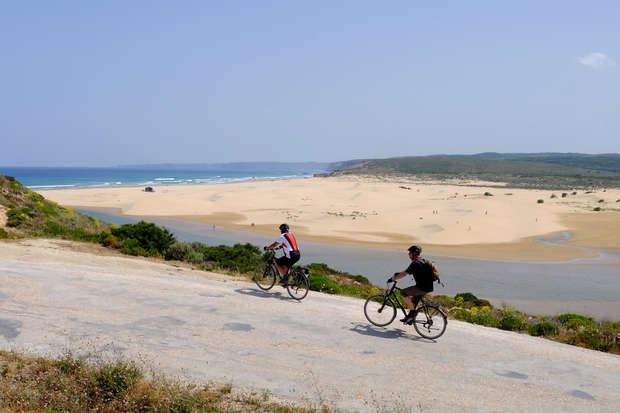 Two cyclists ride along a paved road near a sandy beach. The coastal landscape features a distant ocean, grassy patches, and a clear blue sky above.