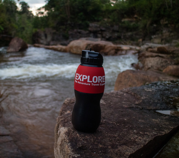 A black and red water bottle labeled "EXPLORE The Adventure Travel Experts" rests on a rock by a flowing river, surrounded by lush greenery and rocky terrain.
