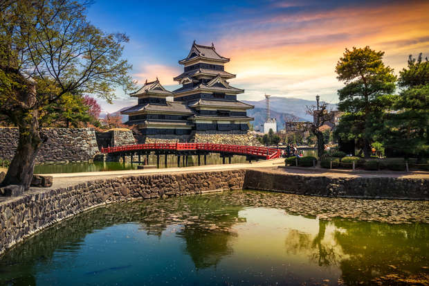 A traditional Japanese castle with tiered roofs stands prominently, reflecting in a tranquil pond, connected by a red arched bridge, surrounded by lush trees under a colorful sunset sky.