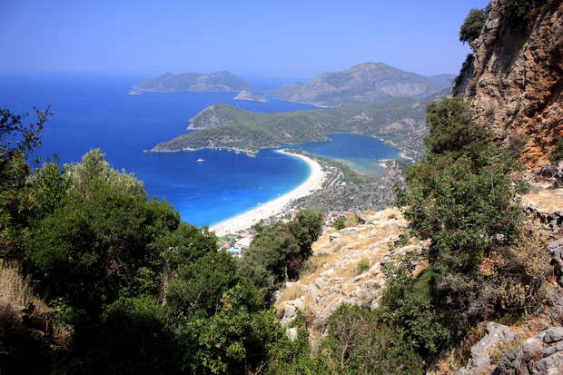 A curved sandy beach stretches between lush green foliage and clear blue waters, with distant mountains under a bright, cloudless sky, viewed from a rocky vantage point.