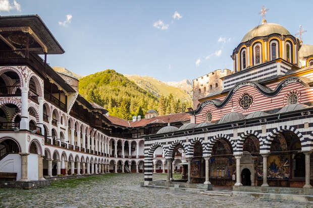 Monastery building features striped arches and domes, adjacent to an open courtyard with cobblestone paving. Surrounded by mountainous landscape with green trees under a clear blue sky.