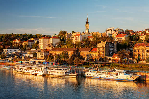 Boats are docked along a serene river, reflecting a vibrant sunset. In the background, a historic cityscape with diverse architecture and a prominent church tower stands on a verdant hill.