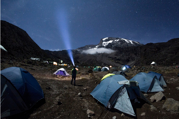 Tents are set up on rocky terrain while a person shines a flashlight skyward. Snow-capped mountain peaks rise in the background under a starry night sky.