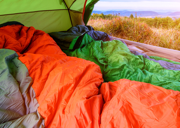 A pair of colorful sleeping bags, orange and green, lie inside a tent. The tent overlooks a scenic outdoor view with grassy fields and distant mountains under a bright sky.