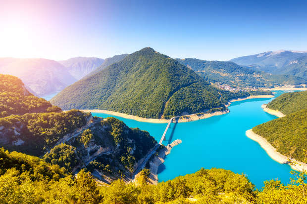 A winding river cuts through lush mountain terrain, with a bridge crossing it. Bright sunlight illuminates the vibrant greenery and vivid blue water under a clear sky.
