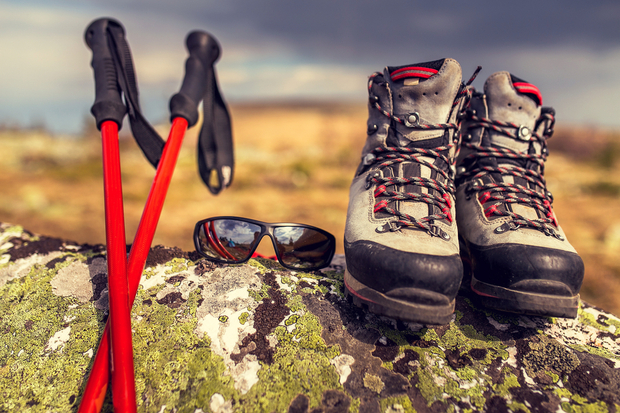 Hiking boots rest on a lichen-covered rock beside red trekking poles and sunglasses, set against a blurred outdoor landscape with a cloudy sky.